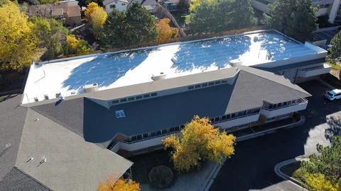 White low-slope roof and gray shingle slopes on a two-story building, with yellow fall trees around it.
