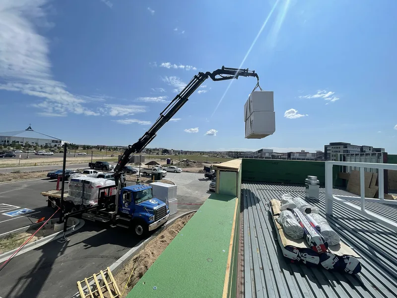 Boom truck lifting a pallet of roofing material up to a commercial metal-deck roof under a blue sky.