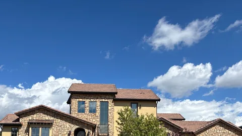 Two-story stone and stucco house with a brown clay tile roof under a bright blue sky with clouds.