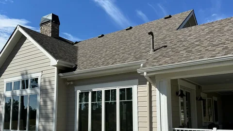 Brown architectural shingle roof with a stone chimney, roof vents and white gutters over tall windows.
