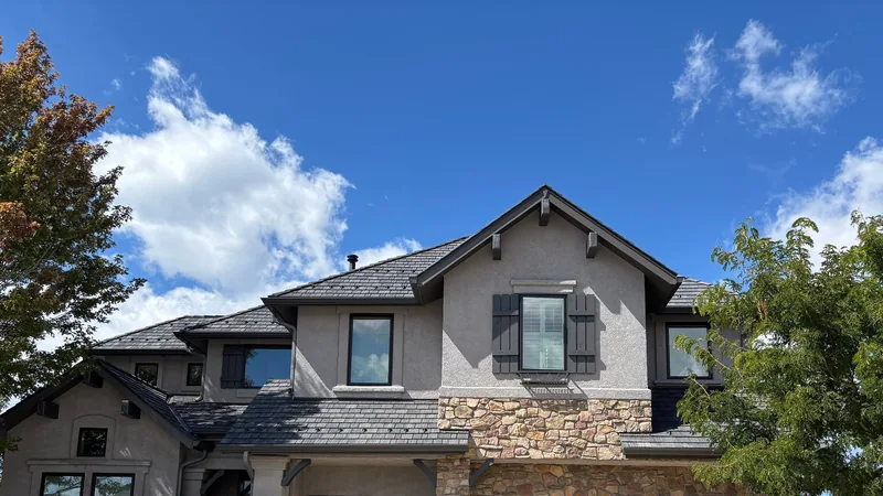 Stucco and stone house with a dark synthetic shake roof under a blue sky with white clouds.