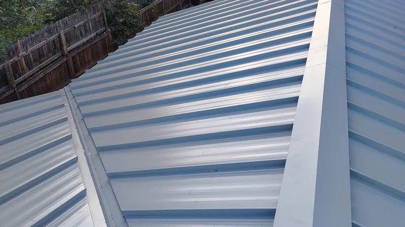 Gray standing-seam metal roof panels meeting at a hip, with a wood fence below.