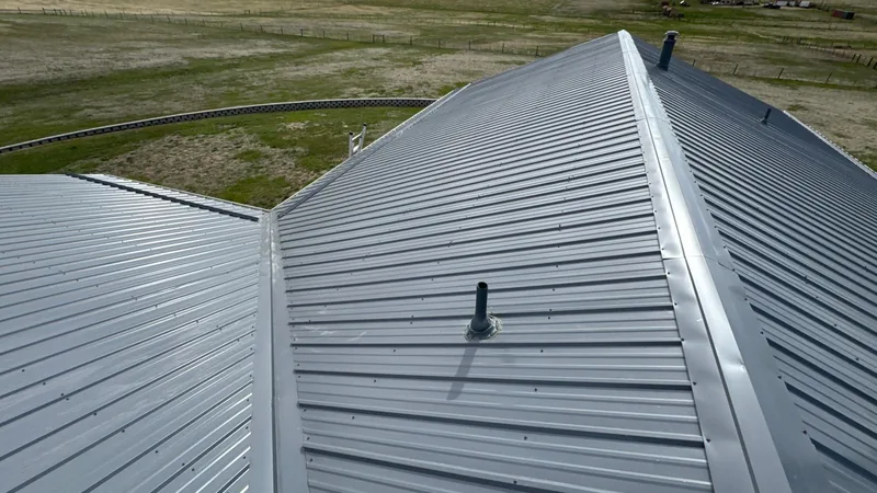 Gray ribbed metal hip roof with a black pipe boot above a wide grassy field.