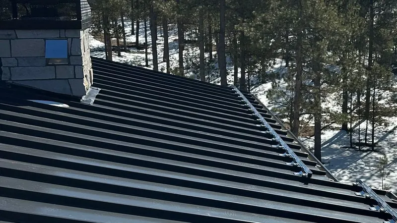 Dark standing-seam metal roof with snow guard rails beside a stone chimney among snowy pines.
