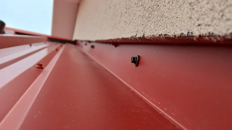 Close view of a red standing-seam metal panel with a fastener against a stucco wall.