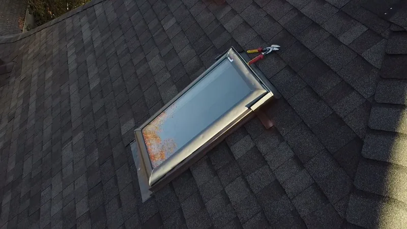 Skylight with metal flashing set into dark gray shingles, with red-handled tin snips beside it.