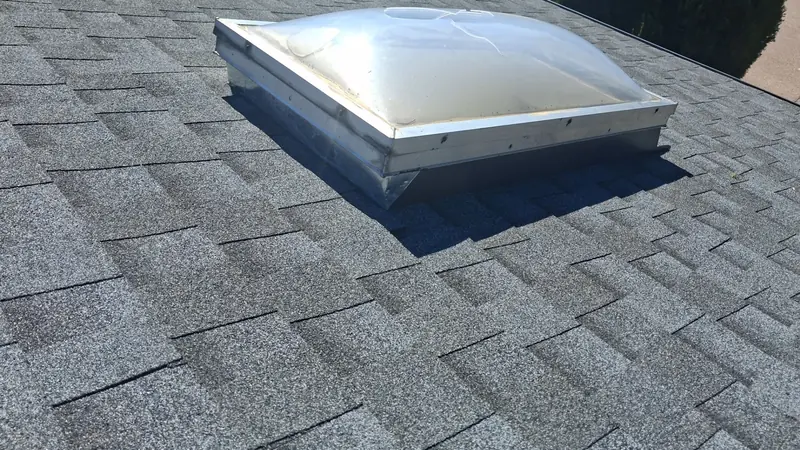 Clear dome skylight on a metal curb set into gray architectural shingles.