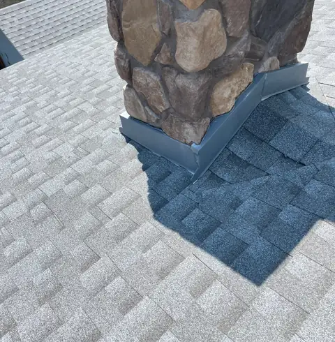 Base of a stacked stone chimney with gray metal flashing on light gray shingles.