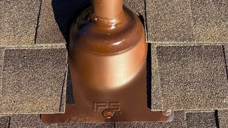 Copper-tone pipe boot flashing set into brown architectural shingles around a vent pipe.