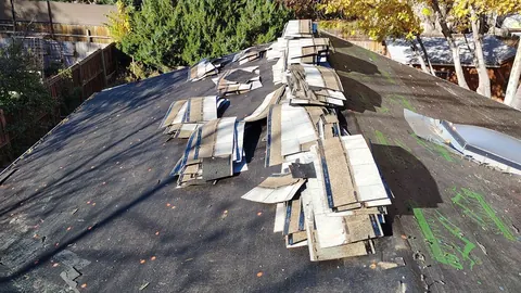Old shingles stripped and piled along a roof ridge above black felt dotted with orange cap nails.